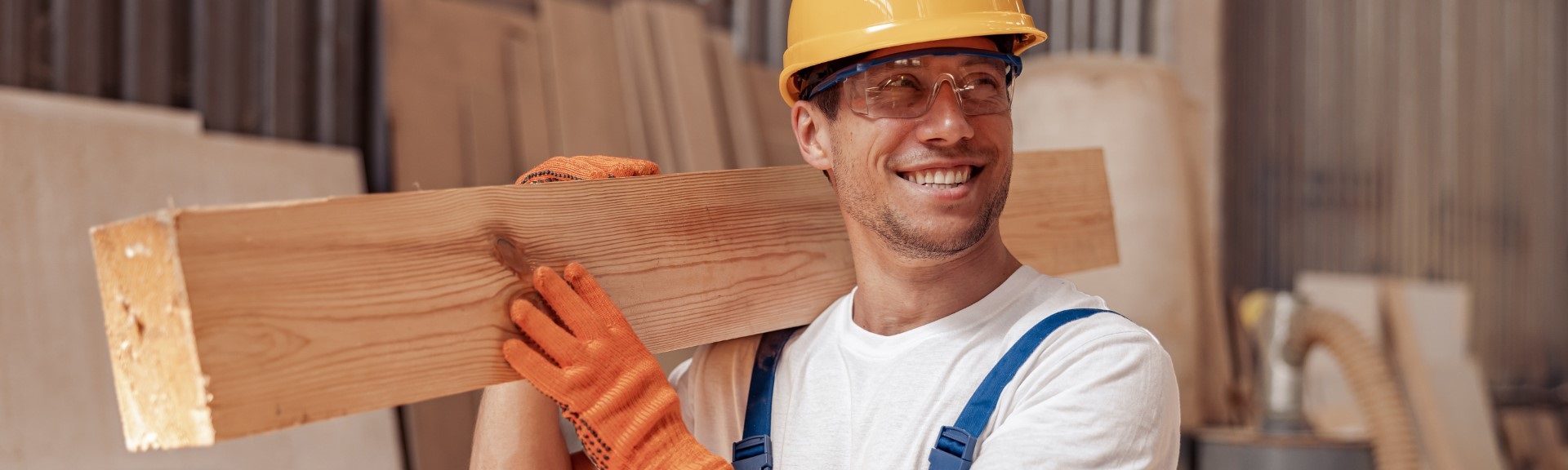 Handsome man builder looking away and smiling while holding wooden plank on his shoulder